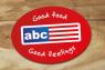 Rood ovalen ABC Restaurants-logo met tekst 'Good food Good feelings' op houten achtergrond.