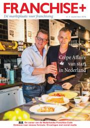 Twee mannen bereiden een pannenkoek in een moderne keuken met menuborden en vers fruit op de achtergrond.