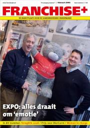 Twee mannen poseren met een grote teddybeer in een galerij met schilderijen op de achtergrond.