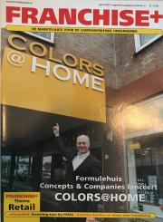 Vooraanzicht van een winkel met de naam COLORS@HOME en een persoon die er met zijn vinger naar wijst.