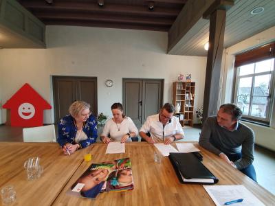 Vier mensen zitten aan een tafel in een kantoor terwijl ze documenten tekenen, met logo van gastouderbureau zichtbaar.
