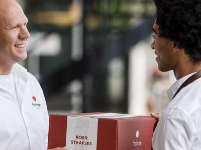 Twee mannen in Fortune-uniform wisselen een rode doos met Fortune-logo en tekst roerstaafjes uit binnenshuis.