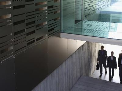 Drie zakelijk geklede personen lopen in een moderne gang met glazen wanden en metalen structuren.