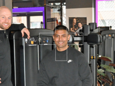 Twee mannen poseren in een sportschool met fitnessapparaten en paarse accenten op de achtergrond.