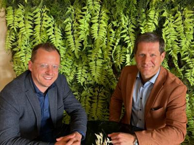 Twee mannen aan een tafel met een vaas droge bloemen, zittend voor een groene plantenmuur.