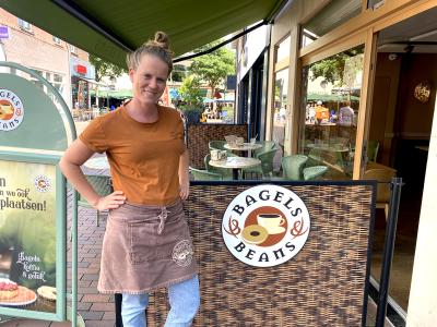 Mandy Herber bij de ingang van haar Bagels & Beans vestiging in Utrecht met logo en terras op de achtergrond.