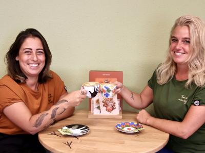 Twee vrouwen proosten met koffiekopjes in een Bagels & Beans vestiging met een menukaart op tafel.
