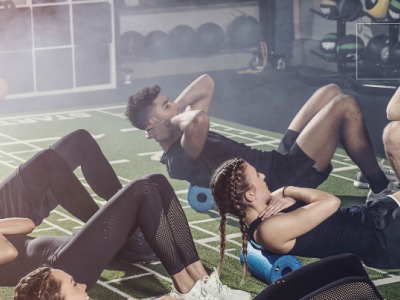 Groep mensen doet buikspieroefeningen op kunstgras onder begeleiding van een coach in een fitnessruimte.