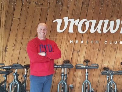 Man in rode trui voor houten wand met het logo van Vrouwfit en een rij spinningfietsen.