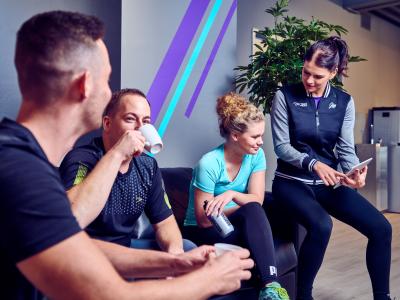 Vier mensen in sportkleding zitten op een bank en praten in een Anytime Fitness-gym.