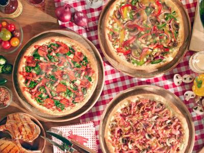 Drie versgebakken pizza's op houten platen met diverse toppings, omringd door ingrediënten en dranken op een geruit tafelk...