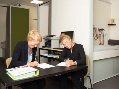 Twee vrouwen schrijven aan een tafel in een studio met fitnessapparatuur op de achtergrond.