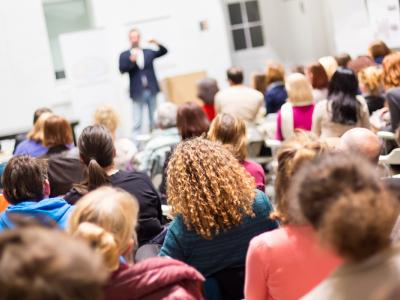 Publiek luistert naar een spreker tijdens een presentatie in een volle zaal.