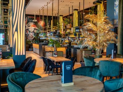 Interieur van een De Beren restaurant met een kleurrijke muur, houten tafels, groene stoelen en sfeervolle verlichting.