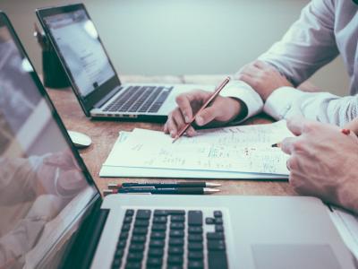 Twee personen overleggen met laptops en papieren op een bureau, met pennen en notities zichtbaar.
