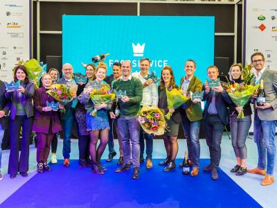 Winnaars poseren met bloemen en awards op het podium van de Foodservice Award met een groot blauw scherm op de achtergrond.