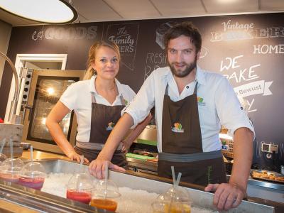 Twee medewerkers in Schorten van Broodje Bewust serveren vers bereide smoothies achter een toonbank.