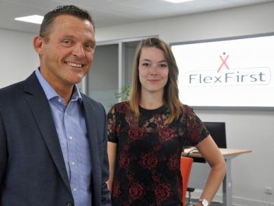 Twee mensen staan in een kantooromgeving naast een FlexFirst-logo op een scherm.