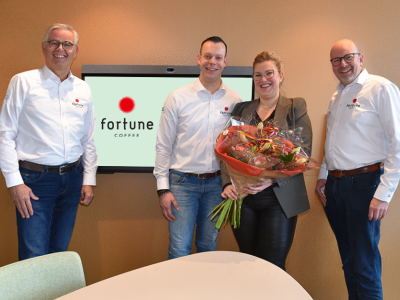 Vier mensen staan glimlachend voor een scherm met Fortune Coffee-logo, waarvan één vrouw een boeket bloemen vasthoudt.