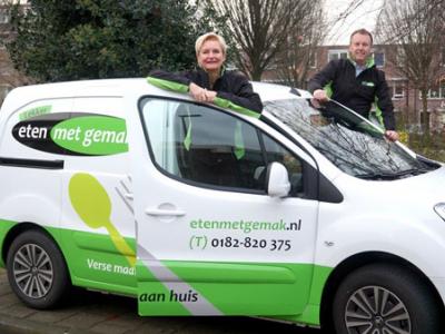 Twee personen poseren naast een bezorgauto van Eten met gemak in een stedelijke omgeving met bomen.