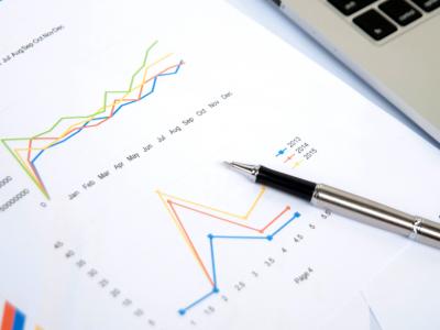 Laptop en pen op een bureau met diverse financiële grafieken en papieren verspreid.