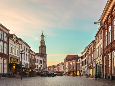 Historische winkelstraat met diverse gevels en een klokkentoren onder een heldere lucht in de avondschemering.