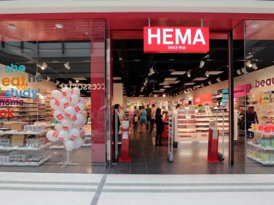 Entree van een HEMA-winkel met rode gevel, winkelend publiek binnen en verschillende producten uitgestald.