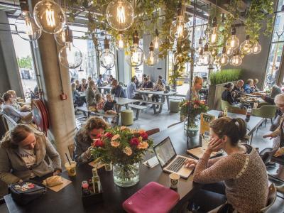 Drukke eetruimte met mensen aan tafels in een moderne setting met hanglampen en planten.