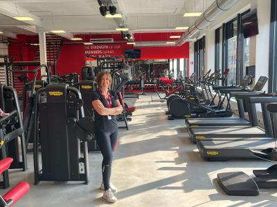 In een goed verlichte gym vol fitnessapparatuur staat een vrouw met gekruiste armen en glimlachend bij krachtmachines.