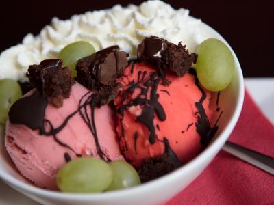 Roomijscoupe met chocoladesaus, slagroom, druiven en brownieblokjes in een witte kom op een rood servet.