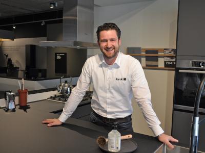 Man in Kvik-werkkleding glimlacht in een moderne keuken naast een fornuis en keukenaccessoires.