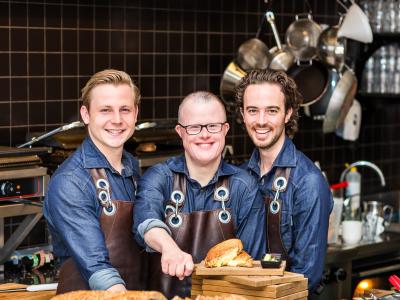 Drie medewerkers in schorten staan glimlachend achter de toonbank met vers gesneden brood en tosti's.