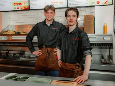 Twee jonge mannen in Kwalitaria-uniform staan in een moderne keuken met frituur en menu's op de achtergrond.