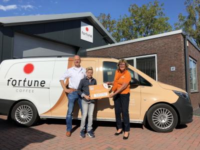 Medewerkers van Fortune Coffee overhandigen een cheque aan KiKa voor het bedrijfsgebouw naast een geparkeerde bedrijfsauto.