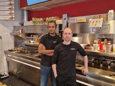 Twee medewerkers staan in een Kwalitaria keuken met een menubord zichtbaar op de achtergrond.