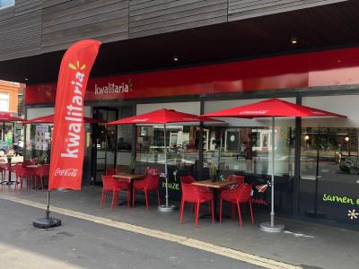 De ingang van een Kwalitaria met rode parasols, tafels en stoelen op het terras en een opvallende reclamezuil.
