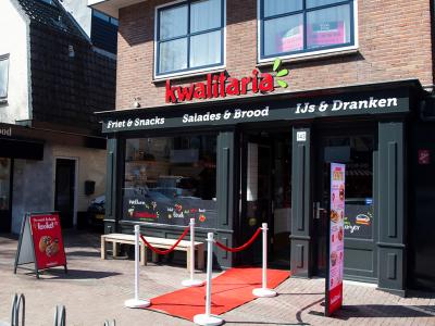 Voorkant van een Kwalitaria-vestiging met rode loper en reclameborden in winkelstraat De Bilt.