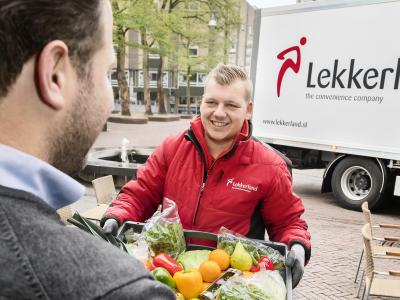 Een medewerker in een rode jas van Lekkerland overhandigt een krat met groenten en fruit aan een klant.