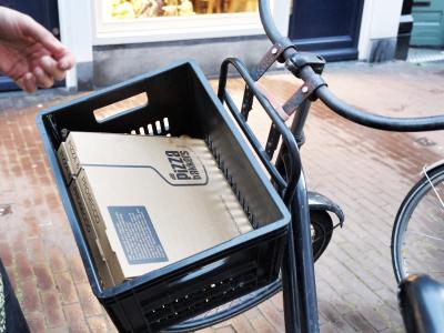 Pizzadozen van De Pizzabakkers in een fietsmand op een straat met winkels op de achtergrond.