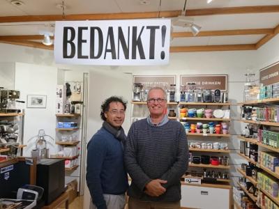 Twee mannen staan in een winkel met een BEDANKT!-bord boven hen, omringd door koffie- en theeproducten.