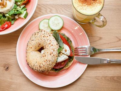 Versierde bagel met groenten en mozzarella op een bord, met bijgerecht en een glas koffie op houten tafel.