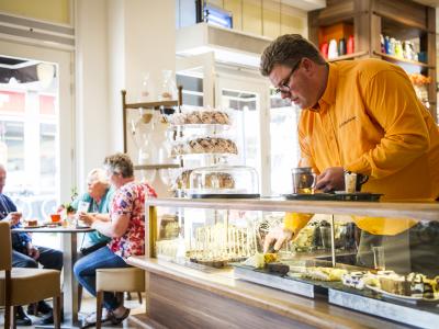 Man in oranje overhemd serveert thee en gebak aan tafel in een gezellige horecazaak.