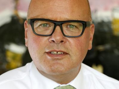 Man met bril en pak poseert voor een kleurrijke, onscherpe achtergrond.