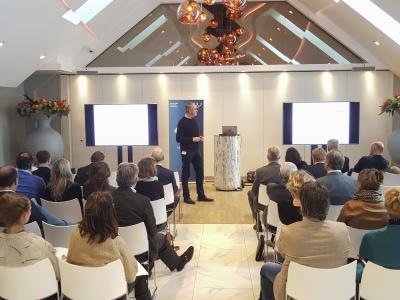 Man presenteert aan publiek in moderne conferentieruimte met schermen en bloemen, gerelateerd aan NFV-bestuur.