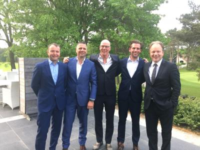 Vijf mannen in nette pakken poseren samen buiten op een terras met groene bomen op de achtergrond.