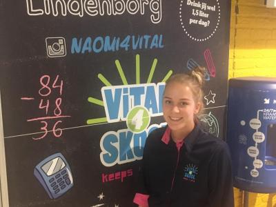 Een jonge vrouw in Vital4Skool-uniform staat voor een kleurrijke muurschildering met educatieve thema's.