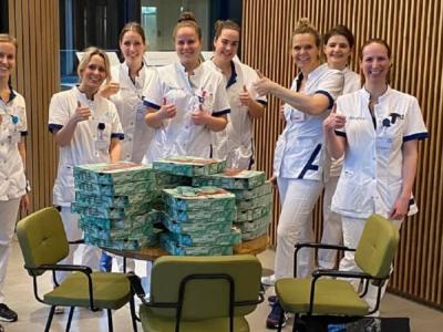 Verpleegkundigen in witte uniformen staan glimlachend rondom een tafel vol stapels pizzadozen in een ziekenhuisomgeving.