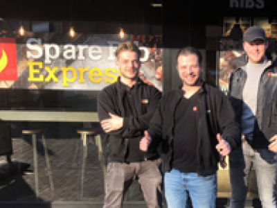 Drie medewerkers poseren voor de Spare Rib Express gevel op een zonnige dag.