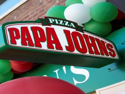 Schuin bord met Papa John's-logo gehangen aan een gevel, omringd door groene en rode ballonnen.