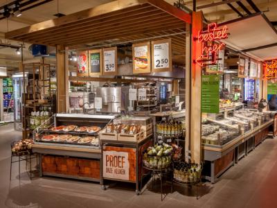 Interieur van een supermarkt met een versafdeling en diverse producten zoals pizza's en broodjes onder neonverlichting.